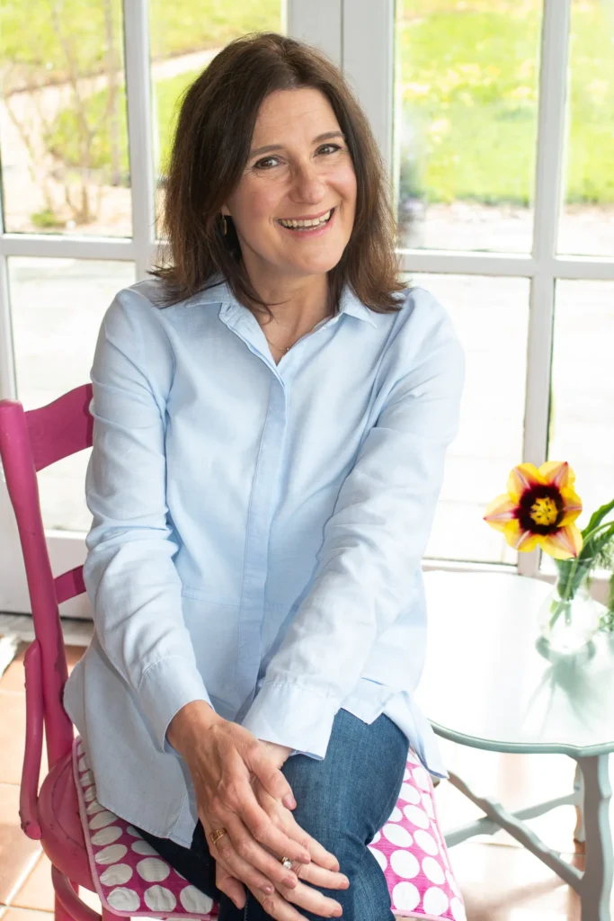 A woman sitting on a pink chair wearing a light blue shirt, with a colorful flower vase on the table beside her, emphasizing creativity and calm organization.